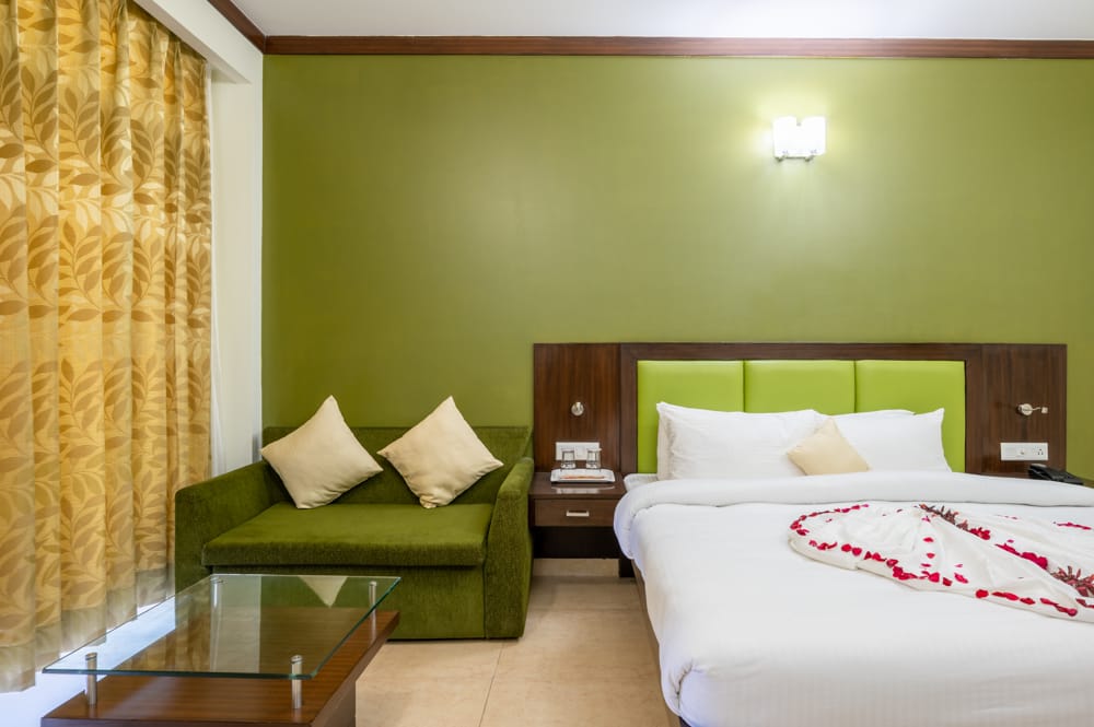 A green room with beautiful floral curtains hanging beside a bed with white sheet placed aside from a green sofa with two cushions